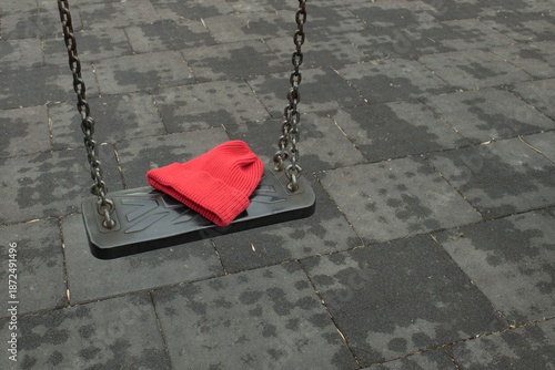 Red knitted beanie hat left on a wet playground swing seat after rain. Concept of lost clothing, childhood, safety reminder, copy space.