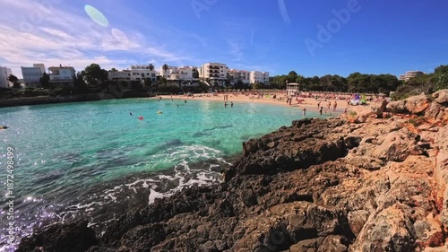 PORTOCOLOM, MALLORCA, SPAIN - OCTOBER 25, 2025: People enjoy the beach and the clear, turquoise water on a sunny day. Ultra-wide pan shot.