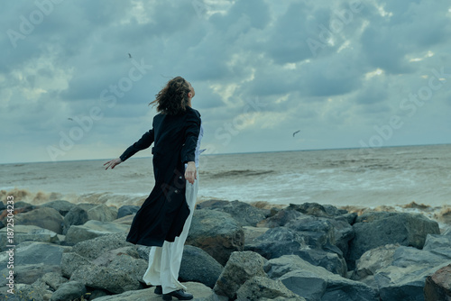 Bild auf Leinwand Woman stands on jagged rocks along the coastline, coat fluttering in the wind, g