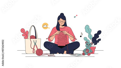 Calm young woman with glasses enjoying her knitting hobby, sitting cross-legged on the floor creating a handmade teddy bear and scarf.