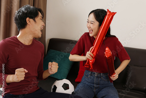 Εκτύπωση καμβά Excited young Asian couple cheering soccer match together on sofa at home holdin