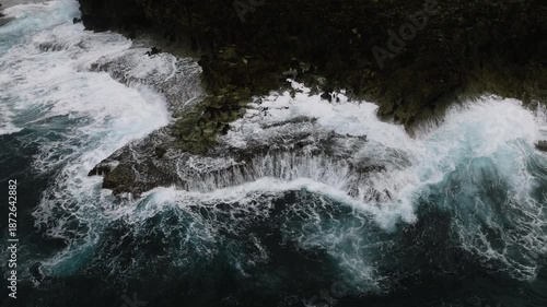 Jagged rocky cliffs are hit by strong white foamy waves under cloudy weather. Siargao, Philippines.