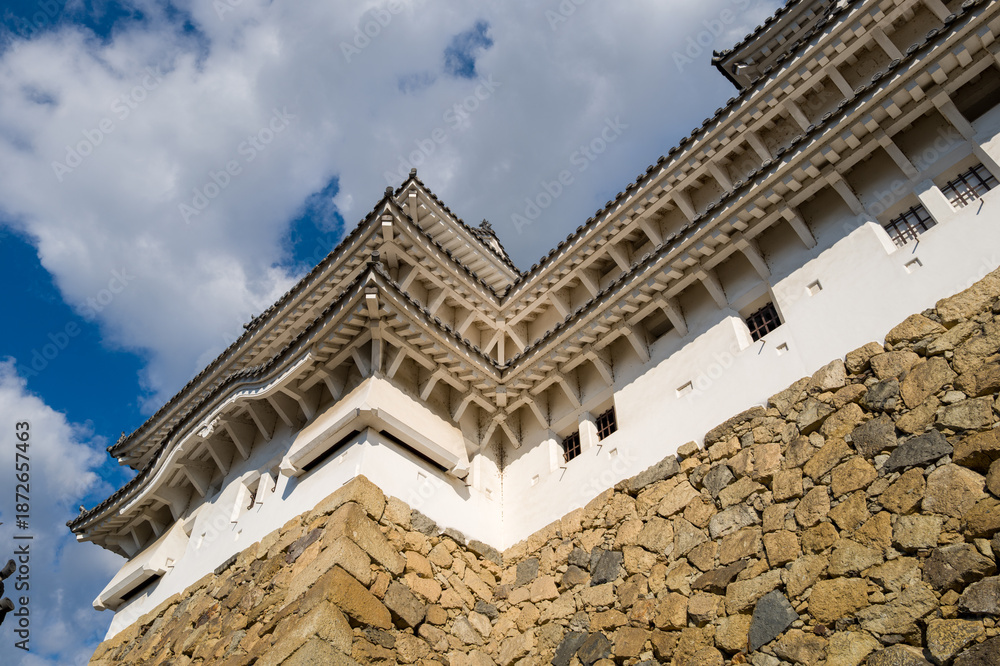 Obraz premium Dramatic upward angle of Himeji Castle's white exterior and layered tiled eaves rising above a textured stone wall, highlighted by sunlight against a partly cloudy sky.