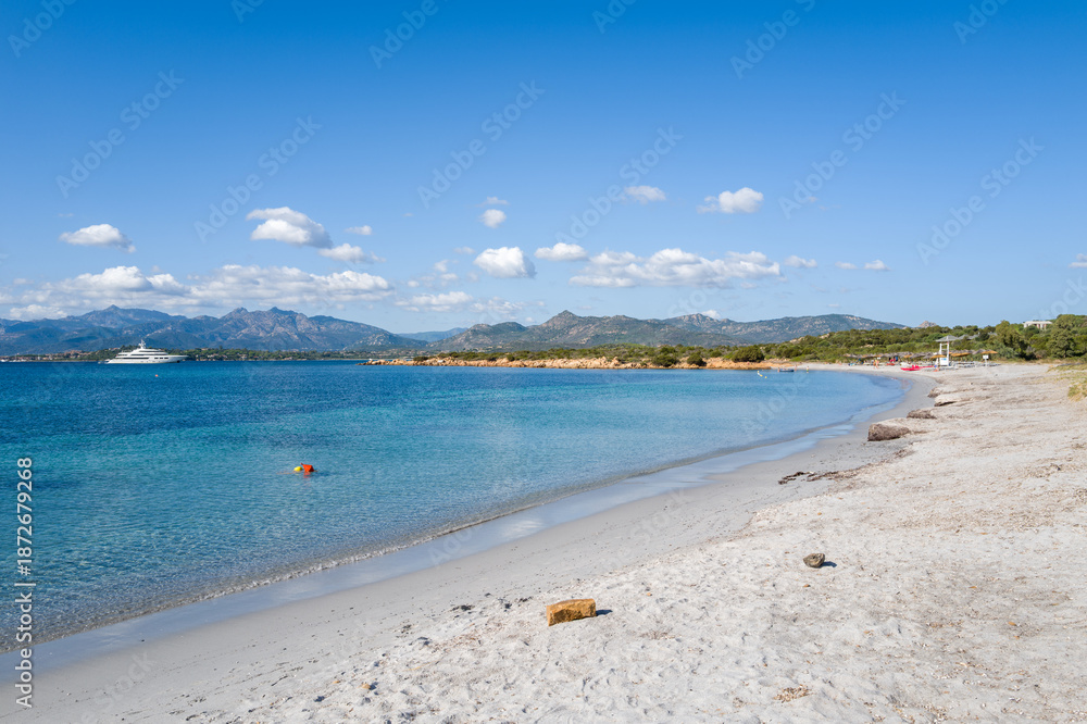 Fototapeta premium Bright white sand curves along the clear turquoise water at Spiaggia la Salinedda in Sardinia, with distant mountains and a yacht under a vivid blue sky dotted with small clouds.