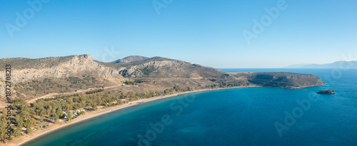Panoramic aerial view of Karathona Beach near Nafplio, Greece, featuring a sweeping sandy shoreline, clear deep blue water, and sunlit rocky hills under a vibrant sky. The landscape is open and