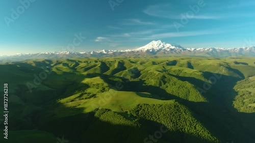 Aerial footage captures a stunning panoramic landscape of the majestic, snow-capped Mount Elbrus, Europe's highest mountain, situated in the Caucasus range. The foreground features vibrant green rolli