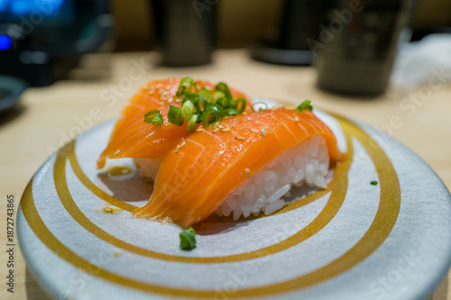 Two pieces of salmon nigiri sushi topped with chopped green onions and sesame seeds are served on a modern silver and gold plate. The fresh fish and glossy rice are highlighted by soft indoor lighting