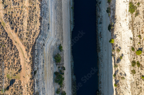 Direct overhead aerial of the Corinth Canal cutting through steep limestone walls, with sparse vegetation and dry, textured earth on both sides under strong sunlight.