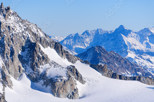 Steep, rugged cliffs dusted with snow rise above the sweeping expanse of Glacier du Geant, with layers of jagged blue mountains fading into the distance under a clear sky.