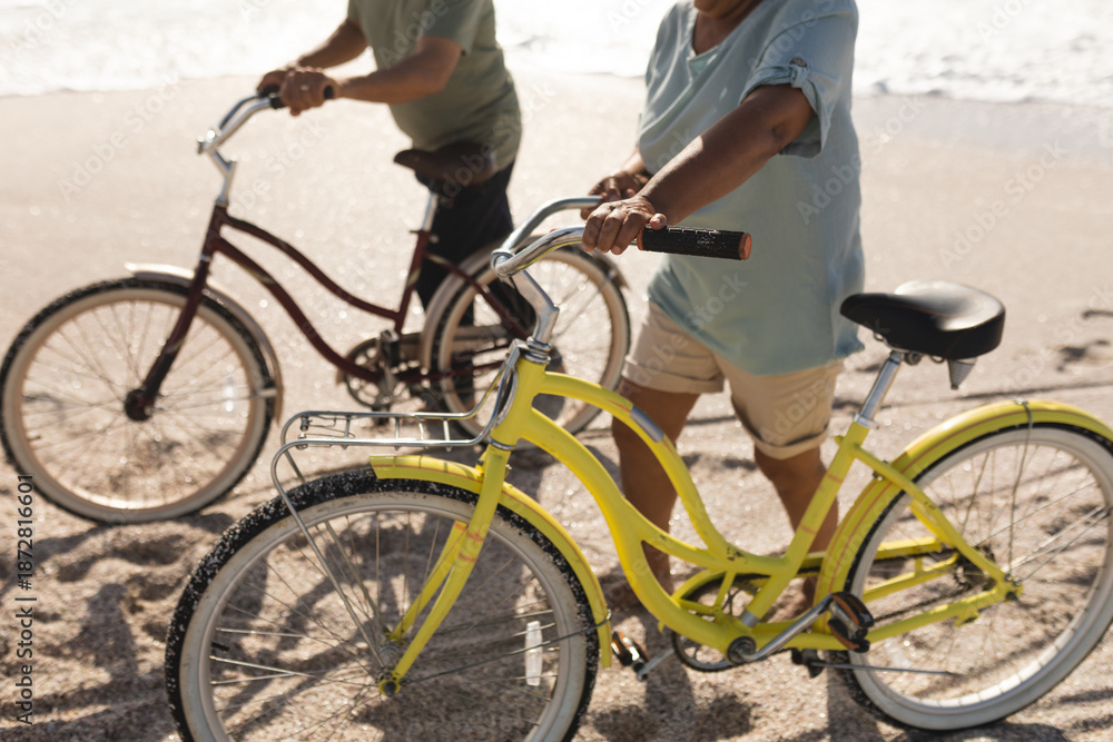 Fototapeta premium Low section of multiracial senior couple walking with bicycles on sand at beach during sunny day