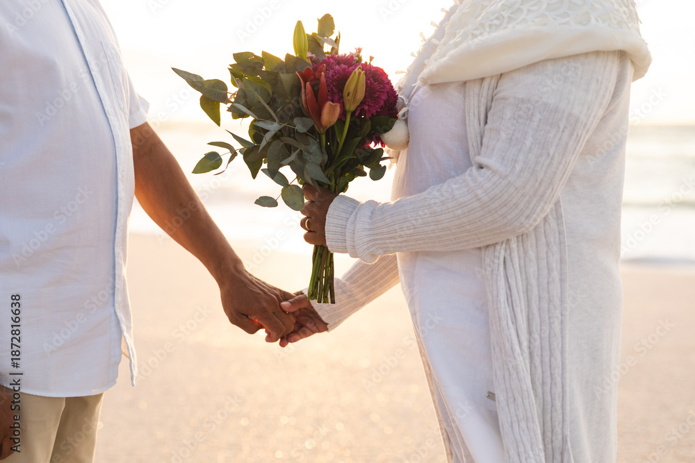 Fototapeta premium Midsection of multiracial senior couple holding hands with bouquet during wedding ceremony at beach