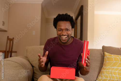 Portrait of cheerful excited african american man opening valentine's present box sitting at home