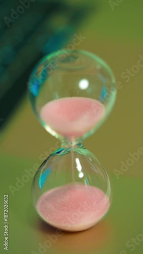 Sand flowing inside hourglass standing on surface. Pink grains running