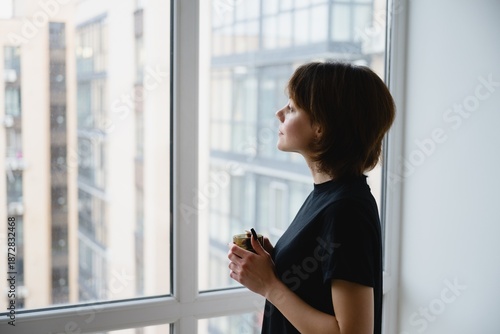 Thoughtful woman holding a cup of coffee while looking out a window at an urban cityscape. Quiet morning moment, mindfulness, lifestyle and calm reflection at home.