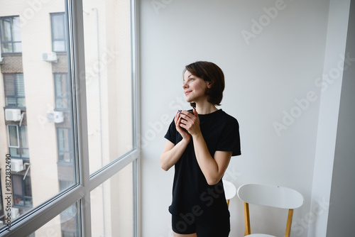 Thoughtful woman holding a cup of coffee while looking out a window at an urban cityscape. Quiet morning moment, mindfulness, lifestyle and calm reflection at home.