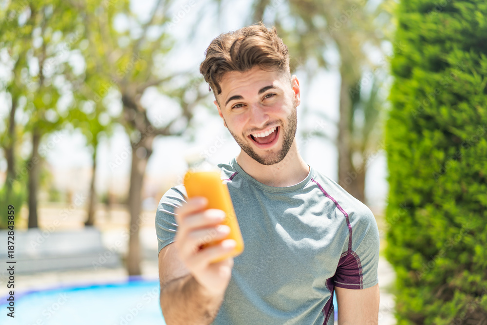Fototapeta premium Young handsome sport man holding an orange juice at outdoors with happy expression