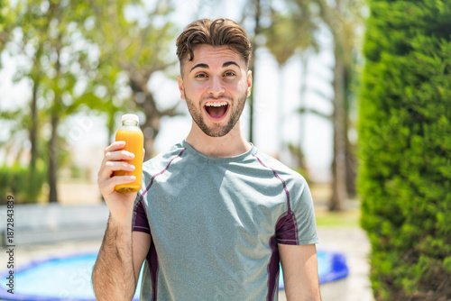 Vászonkép Young handsome sport man holding an orange juice at outdoors with surprise and s