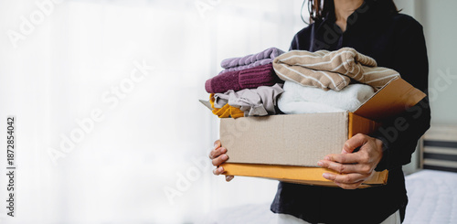 Female hands holding a cardboard box filled with used clothing. Concept of clothes donation, charity, sustainability, recycling, and home decluttering.