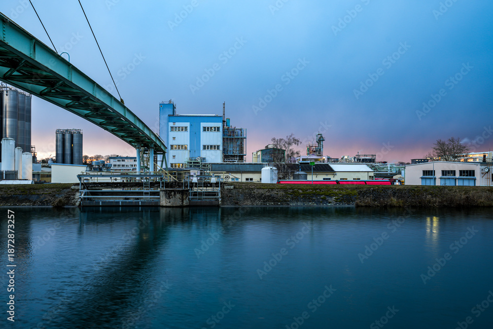 Fototapeta premium The Gersthofen industrial park is a chemical and industrial site with futuristic-looking buildings and silo facilities. The green bridge construction spans the Lech Canal