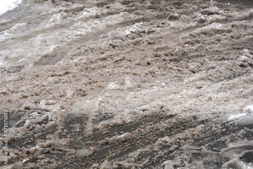 A close up of grey melting slush and dirty snow on a city street with visible tire tracks and messy texture during winter thaw.