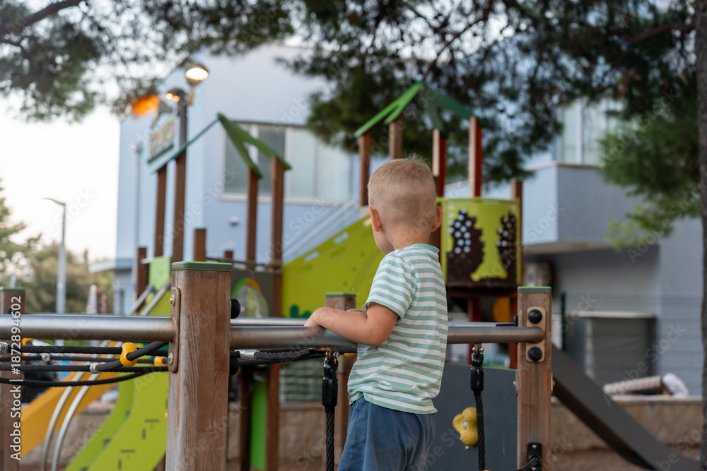 Fototapeta premium Back view of blonde boy in striped shirt standing on playground platform looking at colorful play structures in resort park. Childhood curiosity, outdoor exploration, summer fun and leisure.