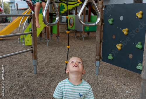 Happy blonde boy looking up at gymnastic rings on playground with climbing wall and slides in summer park. Child anticipation, outdoor play excitement, physical challenge and joyful childhood.
