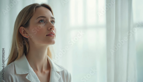 Young woman looks into distance near window. Lady stands in bright room with white curtains. Female model poses in casual shirt with dreamy mood and future hope. Free Download Young woman looks into distance near window. Lady stands in bright room with white curtains. Female model poses in casual shirt with dreamy mood and future hope. - GFXLoad