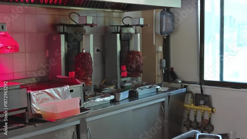 Close up view of doner kebab meat roasting on vertical grills inside small restaurant kitchen with stainless steel work area. Greece. Rhodes.