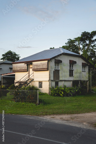 A side-angle view of a modest, elevated house in a lush tropical setting, featuring weathered siding and a classic silver corrugated metal roof. 