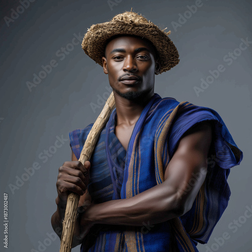 Portrait of muscular man wearing traditional blue clothing and straw hat holding a wooden staff in studio