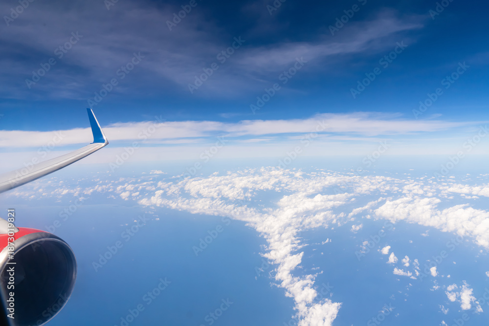 Obraz premium Clouds stretch across the sky as seen from an airplane window. A wing is visible on the right side, with the ocean below reflecting sunlight. This scene shows the vastness of the sky. High quality