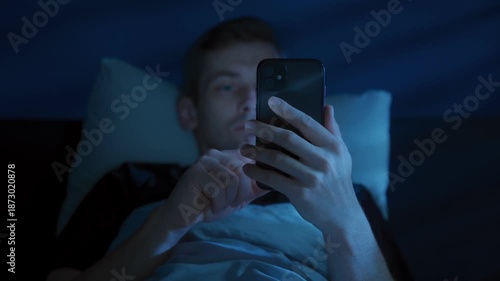 Young man lies in bed holding his mobile phone, scrolling social media at night with his face glowing under blue screen light as insomnia and digital distraction keep him awake. Screen addiction