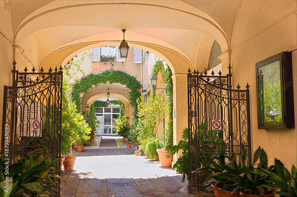 Fototapeta premium Through an archway leading to a courtyard in Rimini. Potted plants line the path, adding greenery to the setting. The area is quiet and inviting for exploration. Rimini, Emilia-Romagna, Italy