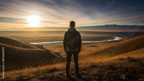 A man with a backpack standing on a hill watching the sun rise over a river valley