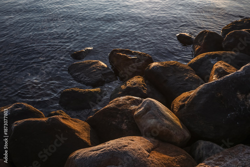 Sunlit coastal rocks and stones surrounded by calm sea water, creating a warm and textured seaside scene at golden hour.
