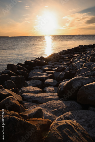 A golden sunset reflecting on calm sea water along a rocky winter shoreline, creating a peaceful and atmospheric coastal landscape.