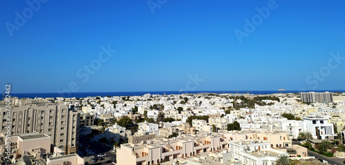 Two storey white buildings of Muscat city center and the Gulf of Oman with Al Fahal island in the distance seen from the rooftop. Panoramic city view