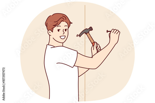 Man with hammer makes repairs in house by hammering nail into wall to hang picture. Young guy is doing repairs to improve interior of apartment or install new shelf to accommodate personal items.