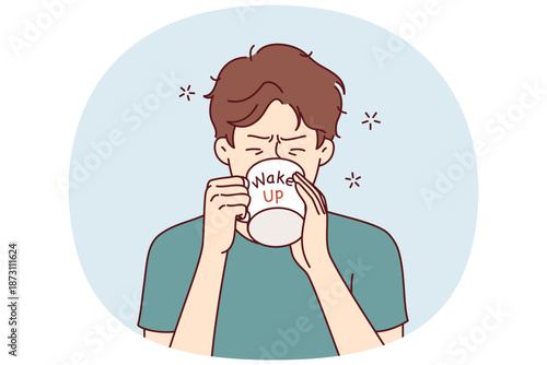 Man drinks coffee from mug with inscription wake up, wanting to cheer up and gain strength before difficult working day. Tired young guy trying to wake up with caffeine and taurine drinks