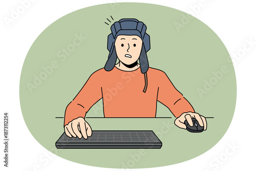 Man gamer plays tanks on computer, sitting at table with keyboard and wears tanker helmet. Guy gamer passionate about video games and virtual tank simulators, makes embarrassed grimace after losing