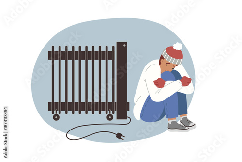 Man is freezing sitting near non-working electric radiator during power outage in house. Guy wrapped in winter clothes is located indoors and is freezing due to lack of heating in apartment.