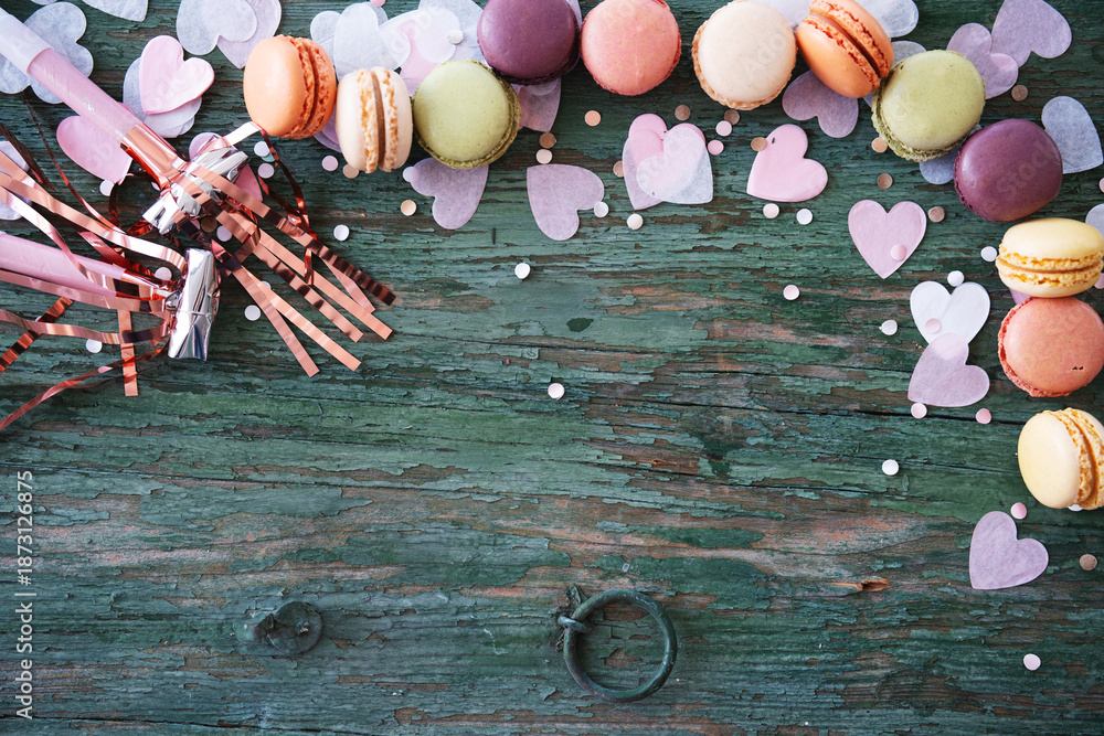 Obraz premium Birthday celebration with delicious macarons surrounded by pink confetti hearts on green shabby chic background. Party and carnival supplies with colorful sweets. Top view with space for text.