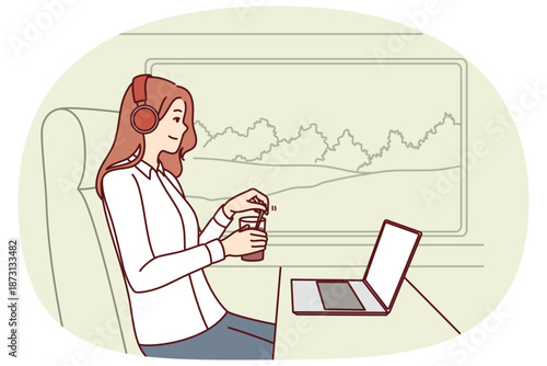 Woman train passenger uses laptop working on trip and drinks coffee sitting near large window in train carriage. Successful girl traveler in headphones sits in train watching movies