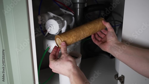 Professional plumber's hands removing a dirty, used cartridge from a reverse osmosis water filter system located under the kitchen sink during routine maintenance for clean drinking water