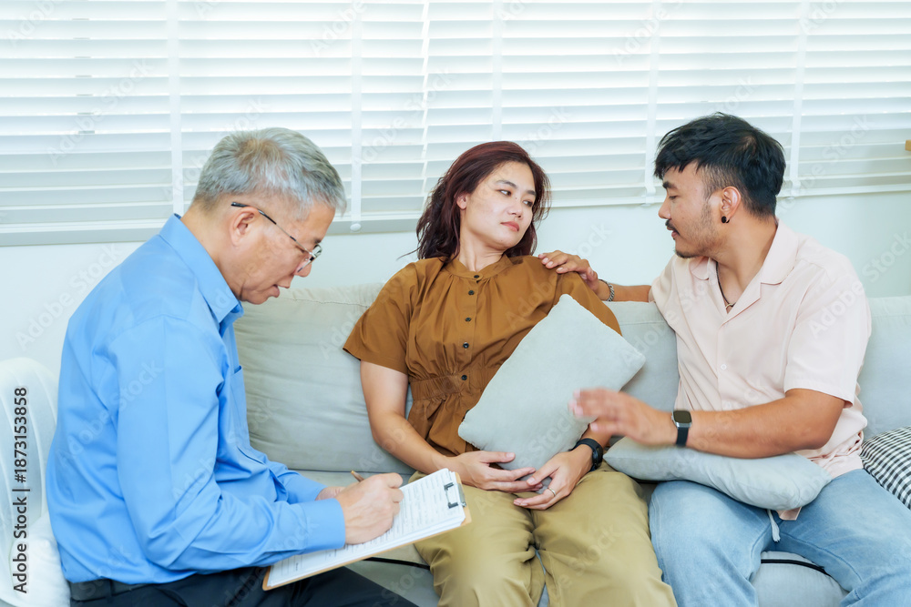Obraz premium Asian couple sitting close during marriage therapy with male psychiatrist writing notes, woman appearing emotional while man comforting gently, relationship counseling, empathy, emotional healing