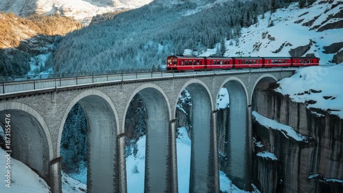Red train crossing high arched bridge in snowy mountain landscape