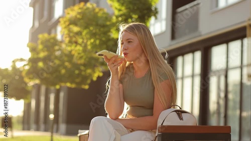 A beautiful young woman with long blonde hair smiles brightly while recording a voice message on her smartphone. 