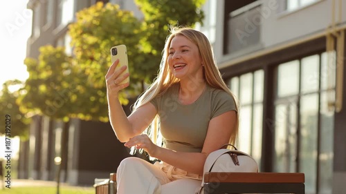 A cheerful young woman with long blonde hair sits on a bench, smiling and waving while engaged in a video call on her smartphone in a bright urban outdoor setting.