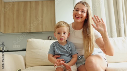 Smiling mother and her adorable son wave cheerfully, greeting the viewer from their cozy home.
