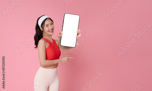 Fitness app promotion smiling Asian woman in activewear holding smartphone mockup and pointing blank screen workout wellness marketing mobile application advertising pink studio background.